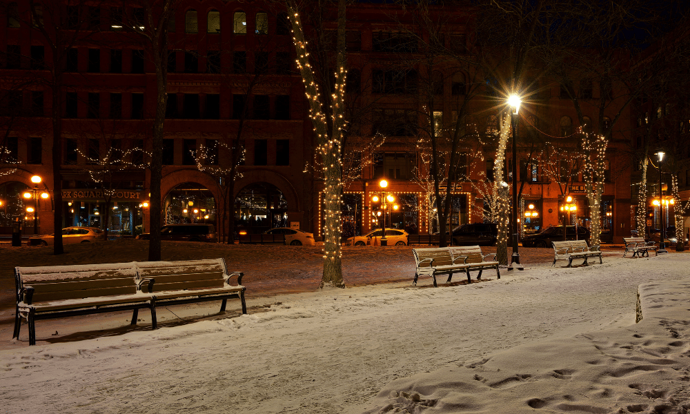 A quiet street is covered in a light snowfall and illuminated by string lights.